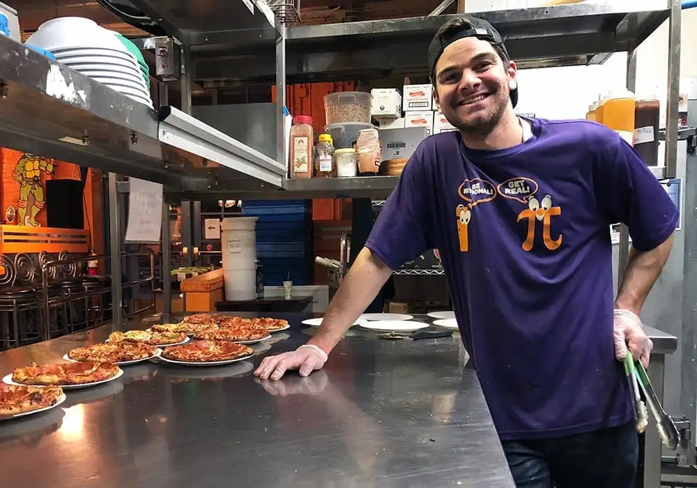 Infinitus Pie employee smiling behind counter with freshly prepared pizzas